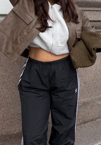 Brązowa skórzana krótka kurtka na szarej bluzie z kapturem na suwak, zestawiona z czarnymi spodniami z elastycznym stanem, ozdobionymi białymi paskami po bokach i logo.