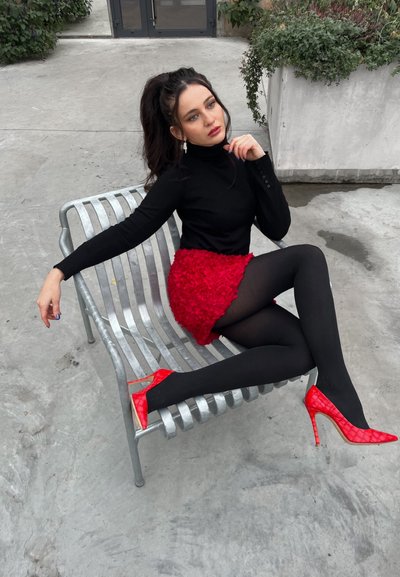 Suéter negro de cuello alto, falda roja texturizada, medias negras, tacones altos rojos de charol. Sentada en un banco de metal, con vegetación de fondo.