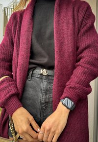 Καρδιά με πλεγμένο πλέγμα σε μπορντό απόχρωση, φορεμένη πάνω από μαύρο καλσόν. Περιλαμβάνει μια μαύρη ζώνη με χρυσό κούμπωμα "RL" και ένα smartwatch.