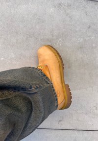 Gamba che indossa un stivale da lavoro in pelle marrone chiaro e jeans di denim scuro, in piedi su un pavimento in cemento grigio liscio.