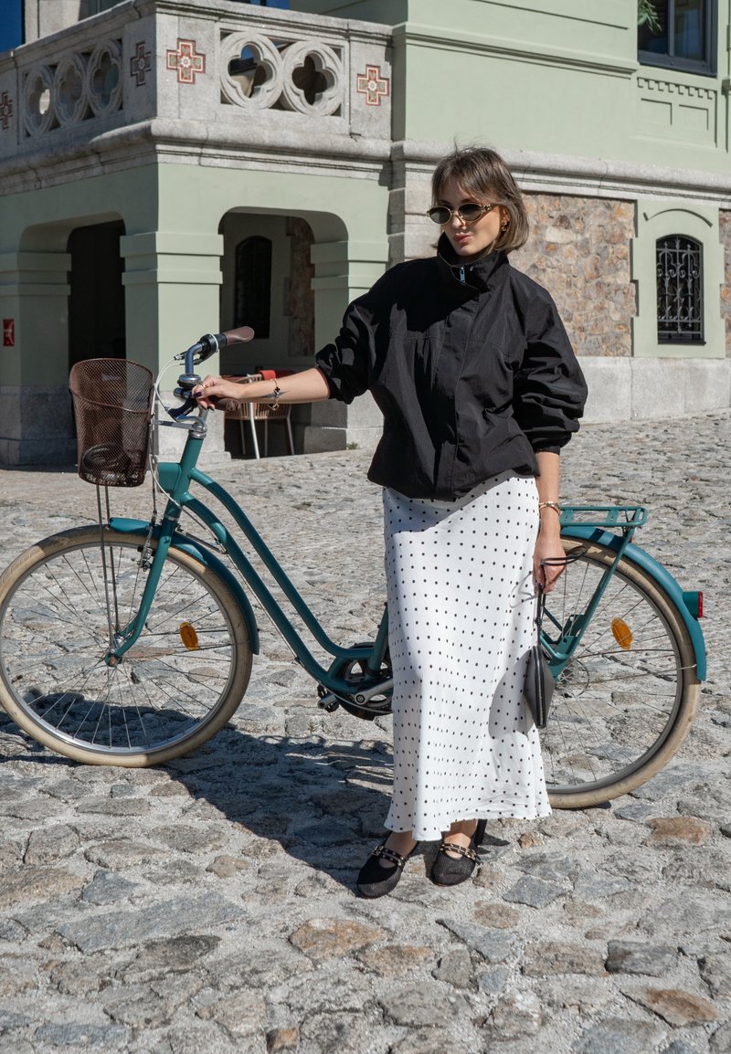 Bicicletta turchese con un cestino di vimini marrone; la ciclista in giacca nera e abito bianco a pois si trova su un sentiero di ciottoli.