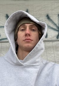 Sweat à capuche gris avec une grande capuche ample, porté par-dessus une chemise de couleur claire. L'arrière-plan présente une surface texturée de couleur claire.