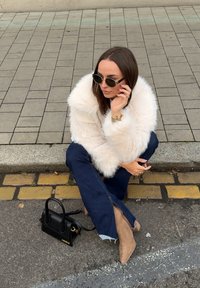 Faux fur-jacka i krämfärg, mörkblå utsvängda jeans med dragkedjor, beige högklackade stövlar och en liten svart handväska.
