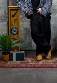 Personne portant une veste texturée rayée, un pantalon noir et des bottes couleur beige se tient sur un tapis à motifs près d'une plante, d'une radio rétro et d'une œuvre d'art musicale encadrée.