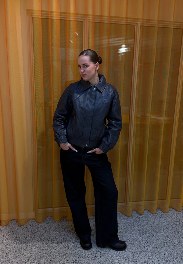 Άτομο με μαλλιά χτενισμένα προς τα πίσω, φορώντας μαύρο δερμάτινο μπουφάν και σκοτεινό παντελόνι, στέκεται με τα χέρια στις τσέπες μπροστά από κίτρινες διαφανείς κουρτίνες.