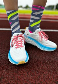 White running shoes with pink laces and a blue sole, paired with gray socks featuring neon pink and green zigzag patterns.