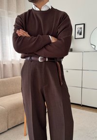 Pull marron porté sur une chemise blanche à col, associé à un pantalon marron taille haute. La tenue est complétée par une ceinture en cuir marron avec une boucle argentée.
