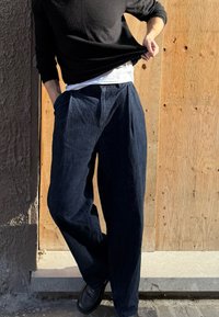 Pantalones de mezclilla holgados de color azul oscuro con pliegues y bolsillos, combinados con una camisa blanca y una camiseta negra de manga larga.