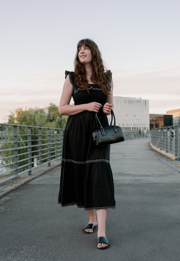 Jeune femme en robe noire tenant un sac à main noir, marchant sur un pont avec des bâtiments modernes et des arbres en arrière-plan.