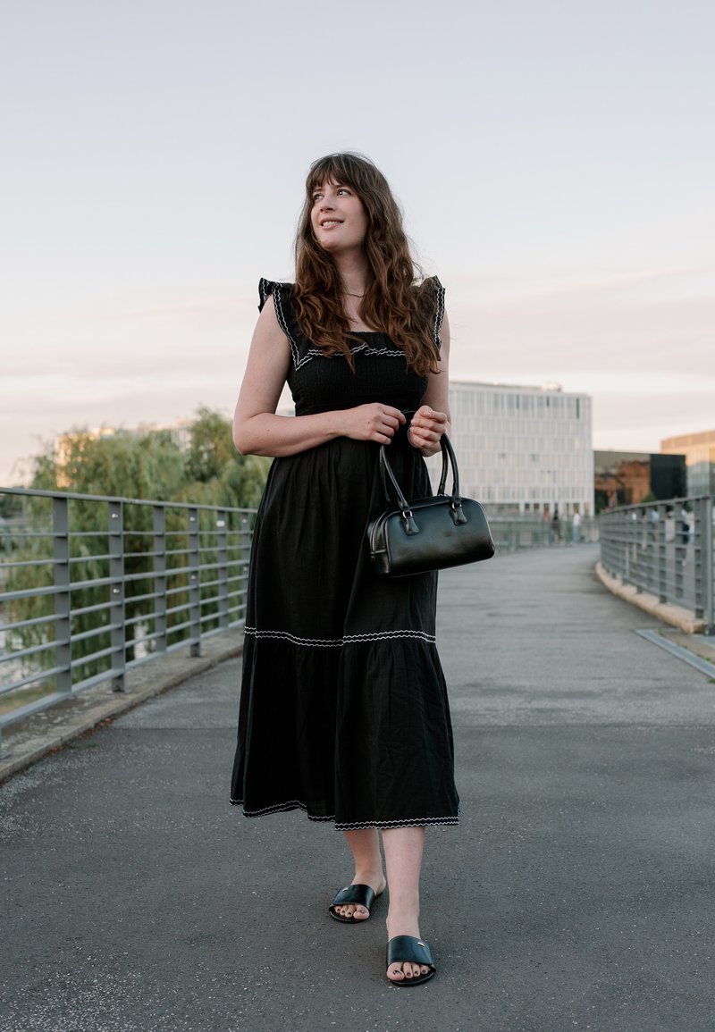 Jeune femme en robe noire tenant un sac à main noir, marchant sur un pont avec des bâtiments modernes et des arbres en arrière-plan.