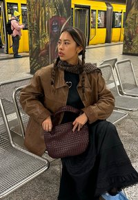 Vrouw met vlechten en een bruine jas zit op een metalen bankje in een metrostation met een gele trein en een orang-oetan muurschildering op de achtergrond.