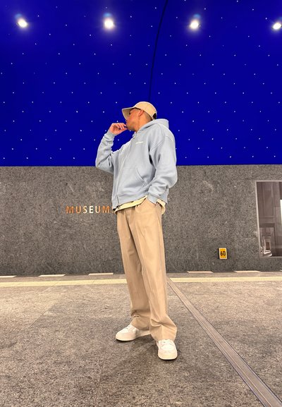 Sudadera azul clara, pantalones beige y zapatillas blancas. De pie en un espacio interior moderno con techo azul y paredes grises.