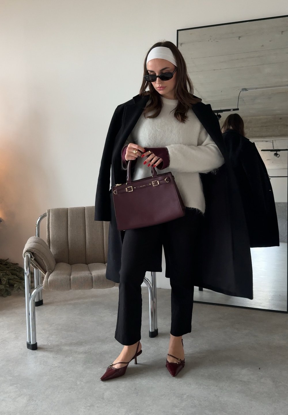 Femme en pantalon noir, talons bordeaux, pull blanc, manteau noir posé sur les épaules, lunettes de soleil et serre-tête, tenant un sac à main bordeaux à l'intérieur.