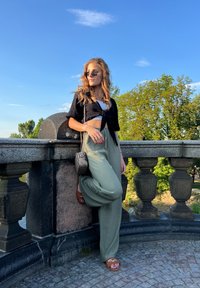 Black cropped top, light green wide-leg pants, brown sandals with chain detail, and small handbag with chain strap. Outdoor setting, blue sky.