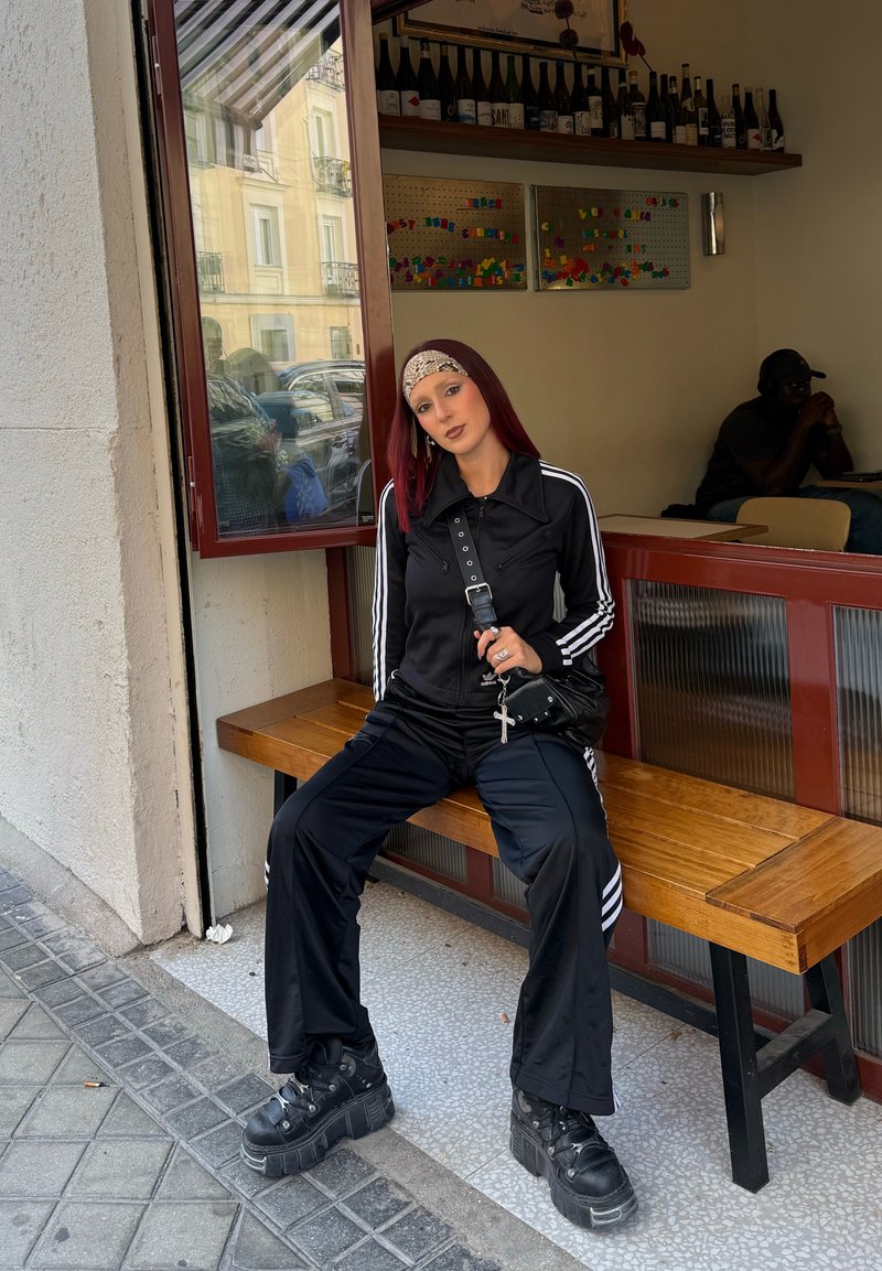Tuta nera con strisce bianche, chunky sneakers neri con platform, lunghi capelli rossi, seduta su una panchina di legno in un ambiente da caffè.