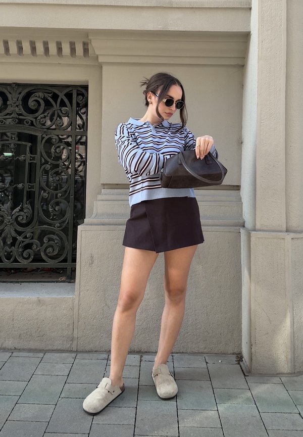 Junge Frau trägt gestreiften Pullover, schwarzen Rock, Sonnenbrille, hält eine schwarze Handtasche, steht an einer beigen Wand und einem verzierten Eisentor.