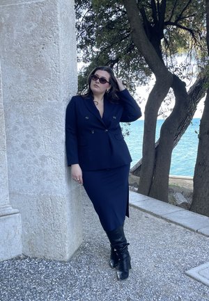 Mujer con blazer y falda azul marino, botas negras y gafas de sol, se apoya contra un pilar de piedra junto a árboles y el mar bajo un cielo despejado.