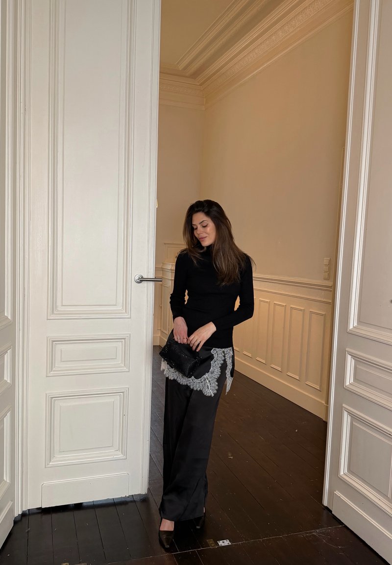 Donna con dolcevita nero e gonna con pizzo bianco che tiene una pochette nera, in piedi in una porta di una stanza con pavimenti in legno e pareti bianche.