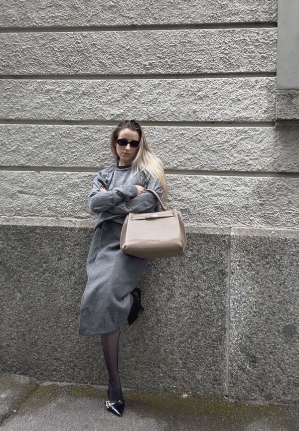 Mujer con atuendo gris y gafas de sol negras, de pie contra una pared de piedra texturizada sosteniendo un bolso beige, con una pierna cruzada sobre la otra.