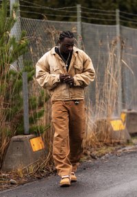 Homme aux cheveux tressés portant une veste et un pantalon beige, marchant sur un chemin pavé à côté d'une clôture métallique et de hautes herbes sèches.