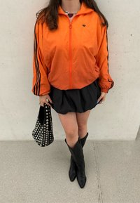 Oranžová bunda na zips s čiernymi pruhmi, čierna rozšírená sukňa, čierne čižmy po kolená a čierna kabelka so cvokmi. Hladká textúra látky.