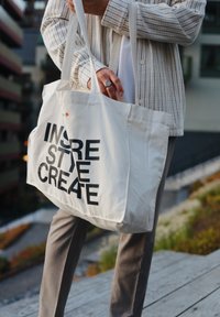 Geantă din pânză albă cu text negru îndrăzneț "INSPIRĂ STIL CREEAZĂ" expus proeminent. Curele ajustabile și design simplu.