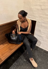 Femme assise sur un banc en bois, regardant dans un grand sac à main noir, portant un gilet beige, un haut noir, un pantalon noir et des chaussures beiges.