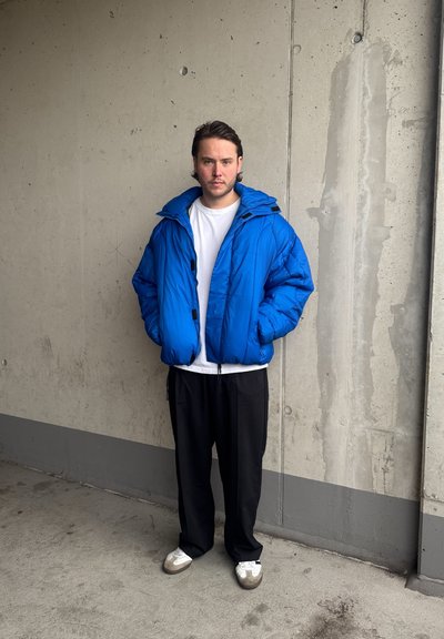 Chaqueta de plumas azul con cremallera frontal y capucha, llevada sobre una camiseta blanca; combinada con pantalones negros y zapatillas blancas. Tejido texturizado, ajuste oversize.