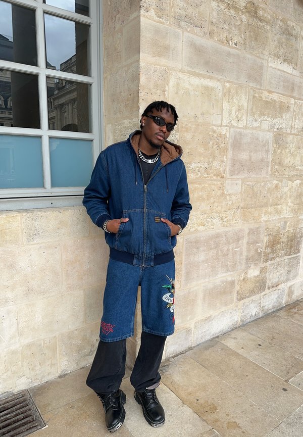Hombre con gafas de sol oscuras, una chaqueta vaquera azul con capucha, pantalones cortos vaqueros sobre pantalones negros y botas negras, de pie contra una pared de piedra.