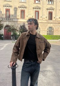 Hnědá bavlněná bunda s límcem a kapsou na zip na hrudi, oblečená přes černou košili. Tmavé džíny doplňují neformální outfit.