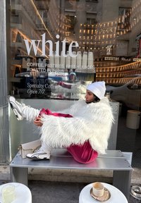 Femme en veste blanche en tissu pelucheux, bonnet et pantalon rose, assise sur un banc en métal à l'extérieur d'un café, avec des tasses de café et des pâtisseries visibles à travers la fenêtre.