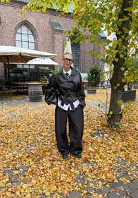 Fekete bőr, oversize dzseki övvel a deréknál, alatta fehér galléros ing, laza sötét nadrágok és fekete táska őszi levelek között.