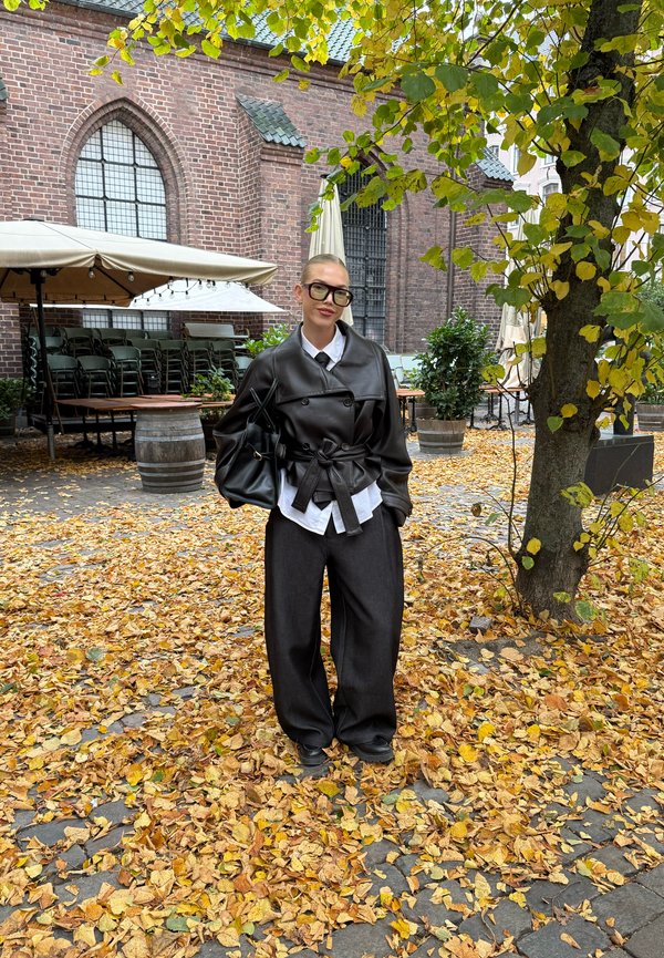Zwarte leren oversized jas met een ceintuur in de taille, een witte blouse eronder, losse donkere broeken en een zwarte tas tussen de herfstbladeren.