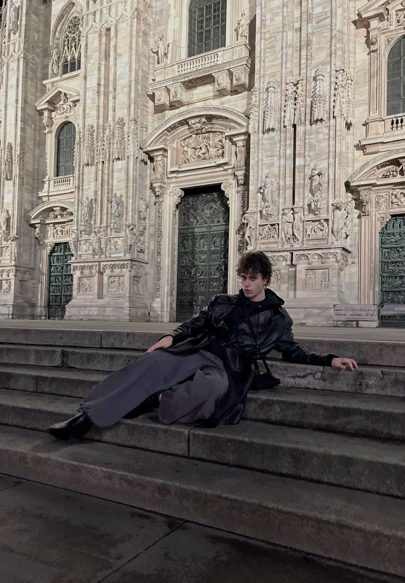 Manteau en cuir noir, pantalon gris large et bottes noires. Personne allongée sur des marches en pierre devant un bâtiment orné et illuminé.