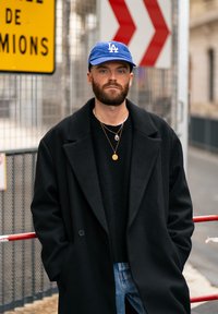 Cappotto lungo nero in lana, maglione nero a maglia, cappellino blu di LA, collane d'oro a strati e jeans azzurri su uno sfondo urbano con segnali di sicurezza.
