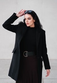 Manteau en laine noire porté sur un pull noir côtelé, associé à une jupe plissée bordeaux. Comprend une ceinture noire avec une boucle ronde dorée et des lunettes de soleil.