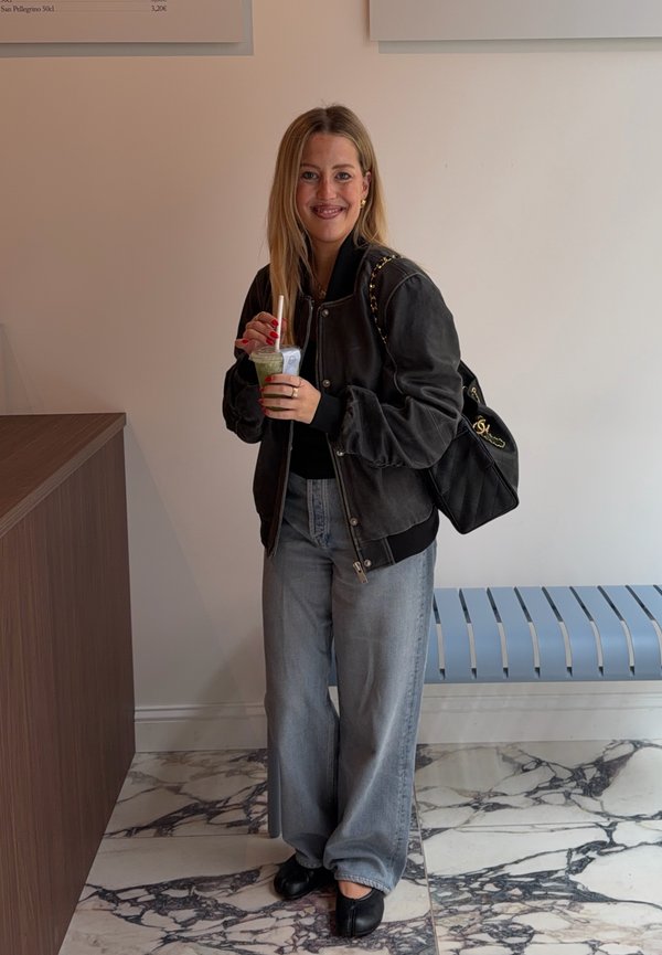 Femme aux longs cheveux blonds portant une veste noire et un jean bleu tient une boisson verte avec une paille dans une pièce minimaliste.