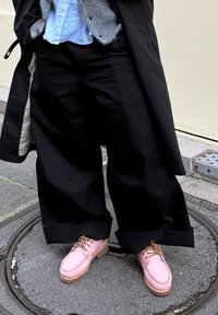 Melnas plata piegriezuma bikses ar šaha raksta oderējumu, pelēks kardigans virs gaiši zilas krekla un rozā apavi ar teksturētu zolīti un brūnām šņorēm.