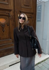 Brązowa dopasowana kurtka z wysokim kołnierzem, w połączeniu z przezroczystą szaro-skalową spódnicą. Dodatki to czarne okulary przeciwsłoneczne i czarna torebka.