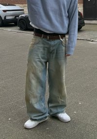 Άτομο που στέκεται στον δρόμο φορώντας φαρδιά ξεθωριασμένα μπλε τζιν, λευκά αθλητικά παπούτσια, μπλε πουλόβερ και καφέ ζώνη με αλυσίδα.