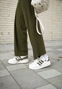 Λευκά αθλητικά παπούτσια με ελαιόχρωμες λεπτομέρειες και σχέδιο με τρεις ρίγες. Πράσινα ριγωτά παντελόνια με ραβδώσεις. Το φόντο έχει ανοιχτόχρωμες τούβλινες πλάκες.