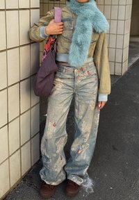 Personne portant une veste en jean courte, une écharpe bleue en peluche, un jean large à motifs, tenant un sac fourre-tout violet, debout près d'un mur carrelé.