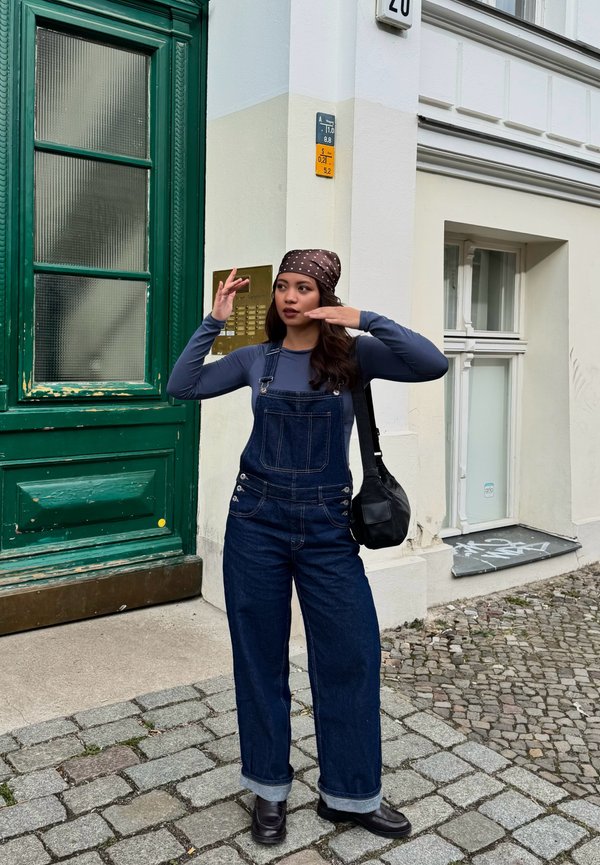 Junge Frau mit braunem Kopftuch und Jeanslatzhose steht auf einer Kopfsteinpflasterstraße und gestikuliert mit beiden Händen nahe ihrem Gesicht.