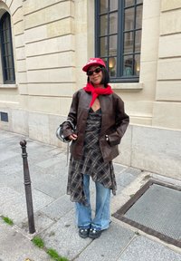 Hnědá kožená bunda přes černé a béžové kostkované šaty, modré široké džíny, červený šátek, červená čepice a černé boty se hvězdami.