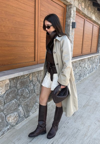 Mujer con gabardina beige, falda blanca, botas negras y gafas de sol caminando junto a un muro de piedra con persianas de madera, sosteniendo un bolso negro tejido.