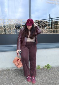 Dunkelrote Lederjacke, beige Pullover, dunkelrote Cordhosen und burgunderrote Turnschuhe. Hält eine pinkfarbene Handtasche und ein Telefon. Strukturierter Hintergrund.