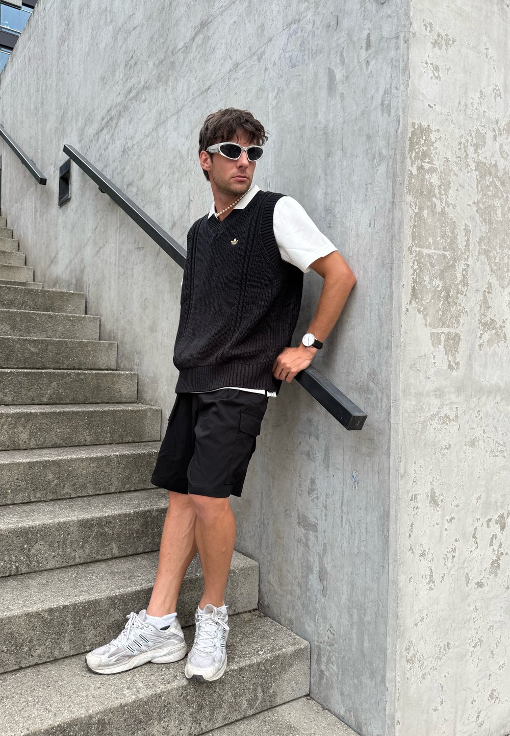 Jovem com óculos de sol, camiseta branca, colete de malha preto, calções pretos e sapatilhas brancas, encostado a uma parede de betão texturizada junto às escadas.