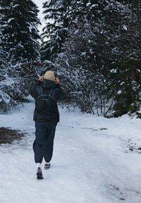 Inimene, kes kannab musta veekindlat jope ja halli pükse, kõnnib lumega kaetud rajal, mida ümbritsevad igihaljad puud.