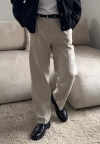 Άτομο με μπεζ παντελόνι, μαύρη ζώνη, μαύρα παπούτσια και μαύρο μπουφάν, που στέκεται σε υφασμένο χαλί κοντά σε έναν μπεζ καναπέ.
