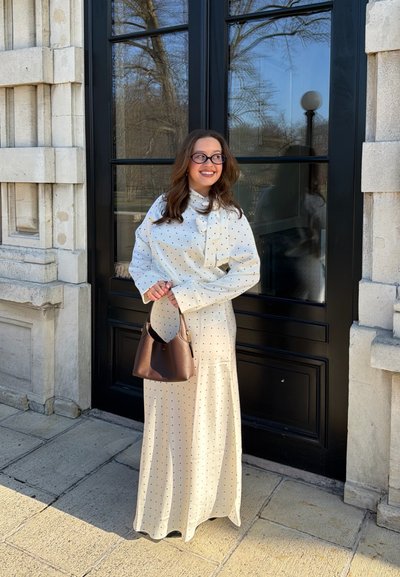 Mujer sonriente de pie al aire libre frente a una puerta negra, vistiendo un vestido blanco con lunares y sosteniendo un bolso marrón.